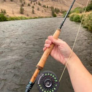Hand holding a fly fishing rod and reel in front of a river