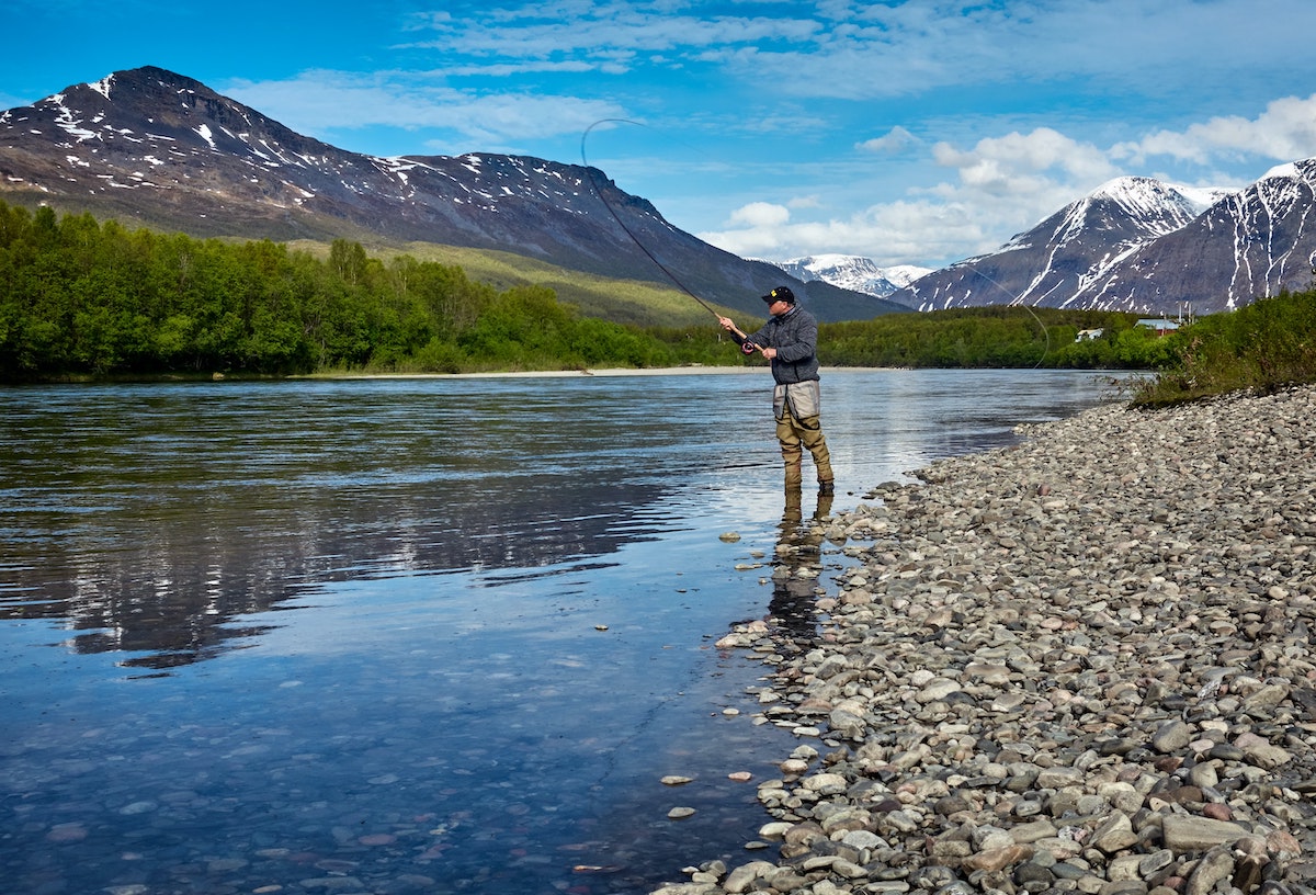 Man standing on the edge of a river fly fishing with mountains in the background